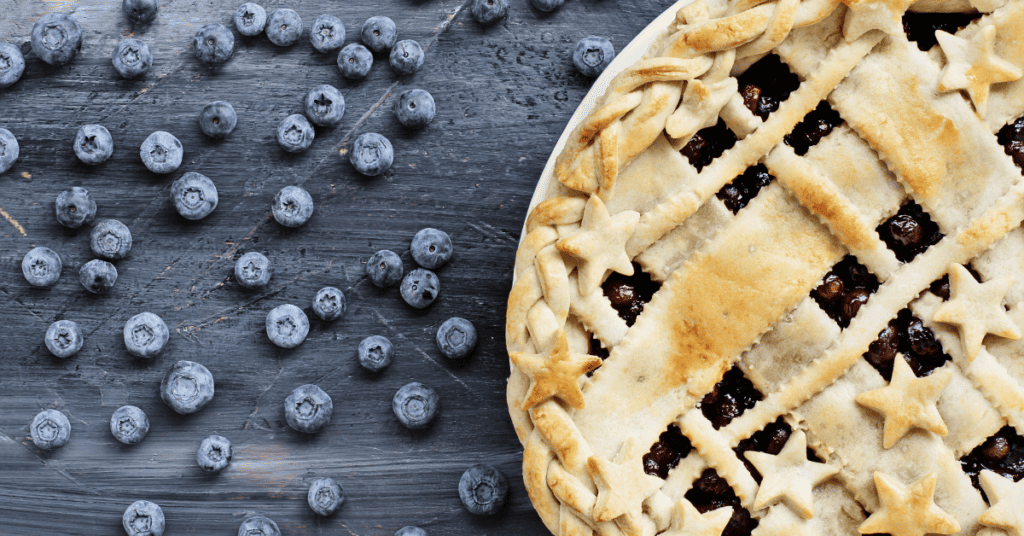 blueberry pie with star lattice crust on top, sitting on countertop with blueberries surrounding it